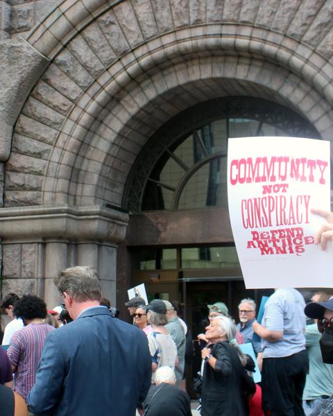 Around fifty people stand mostly backs to the camera facing a stone courthouse with a giant arch door, one holding up a Community Not Conspiracy: Defend Anti-ICE MN15 sign.