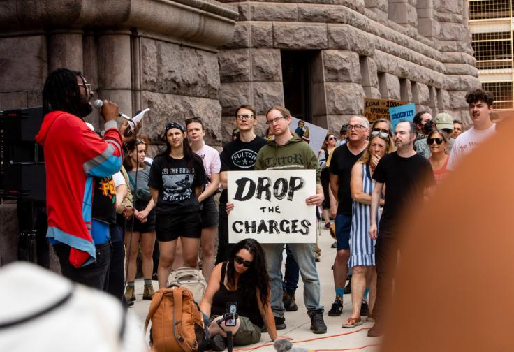 A man speaks energetically into a microphone and another holds a large 'Drop the Charges' sign as dozens of people gather to rally in support of the 15 Minnesotans indicted for conspiracy to impede or injure a federal agent outside of the Diane E. Murphy federal courthouse during their arraignment hearing in a photograph by Nicole Neri.