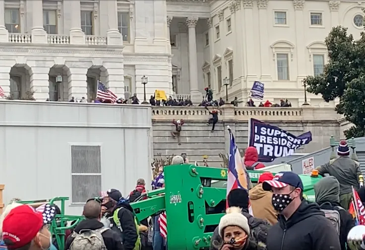 A vacant-looking white man in a black hoodie with the hood down, a Trump 2020 hat, and a US flag facemask stands with a concerned looking white woman also wearing a US flag facemask in front of an "I Support President Trump" banner, while numerous people watch others scale the walls of the Capitol and stand on it, many masked and some carrying Trump signs.