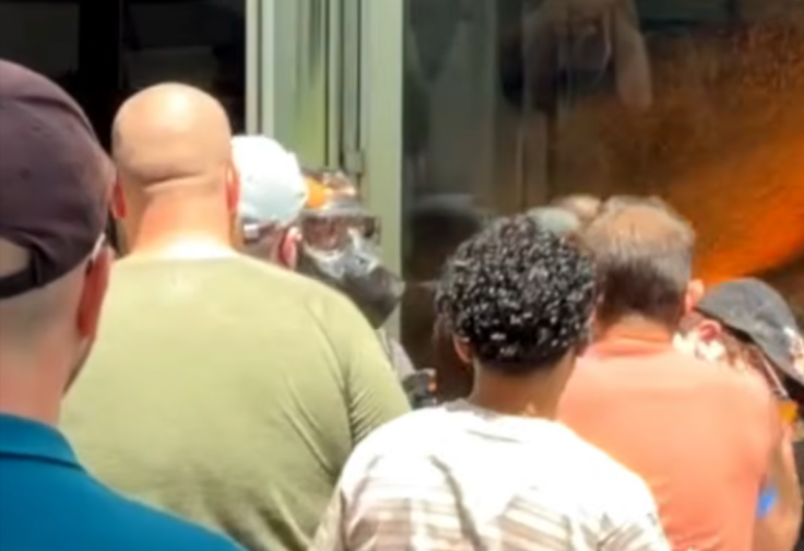 A half-dozen people facing away from the camera at the doors of a courthouse as a police officer in a full gasmask sprays orange chemical at them.