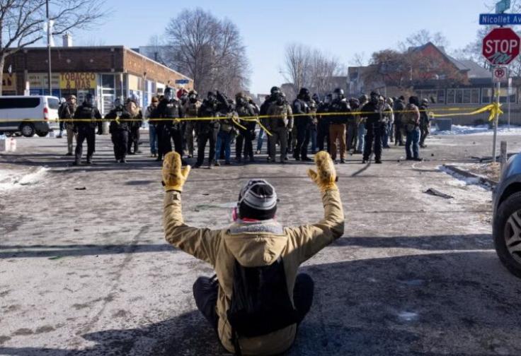 A person wearing a winter hat and yellow gloves sits on a snowy city street, back to the camera, hands up, facing 20 or more armed agents in body armor and riot masks.