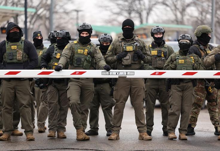 A row of masked, BORDER PATROL FEDERAL AGENT vest and army fatigue wearing apparent white men.