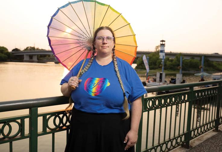 Treasure Thoreson, with long braided pigtails, wearing a blue shirt with a colorful MFE logo and holding a rainbow parasol stands in front of the Mississippi River in Boom Island Park, in a photo by Sarah Whiting.