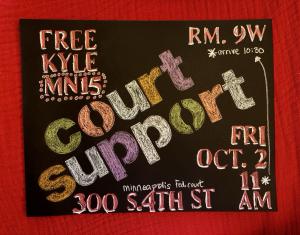 A handmade poster on black paper sits on a red cloth. The text reads as follows: Free Kyle MN15. Court Support; Friday October 2 at 11 a.m. at the Minneapolis Federal Courthouse, 300 S. 4th St; Room 9W. Arrive at 10:30.